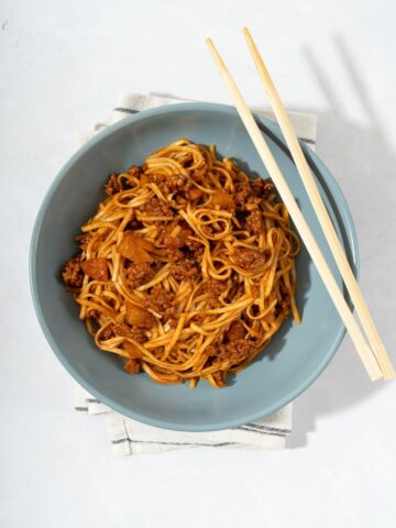 A bowl of kimchi noodles mixed with ground meat and kimchi next to chopsticks.