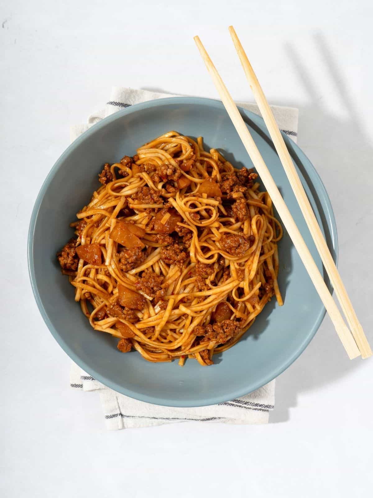 A bowl of kimchi noodles mixed with ground meat and kimchi next to chopsticks.