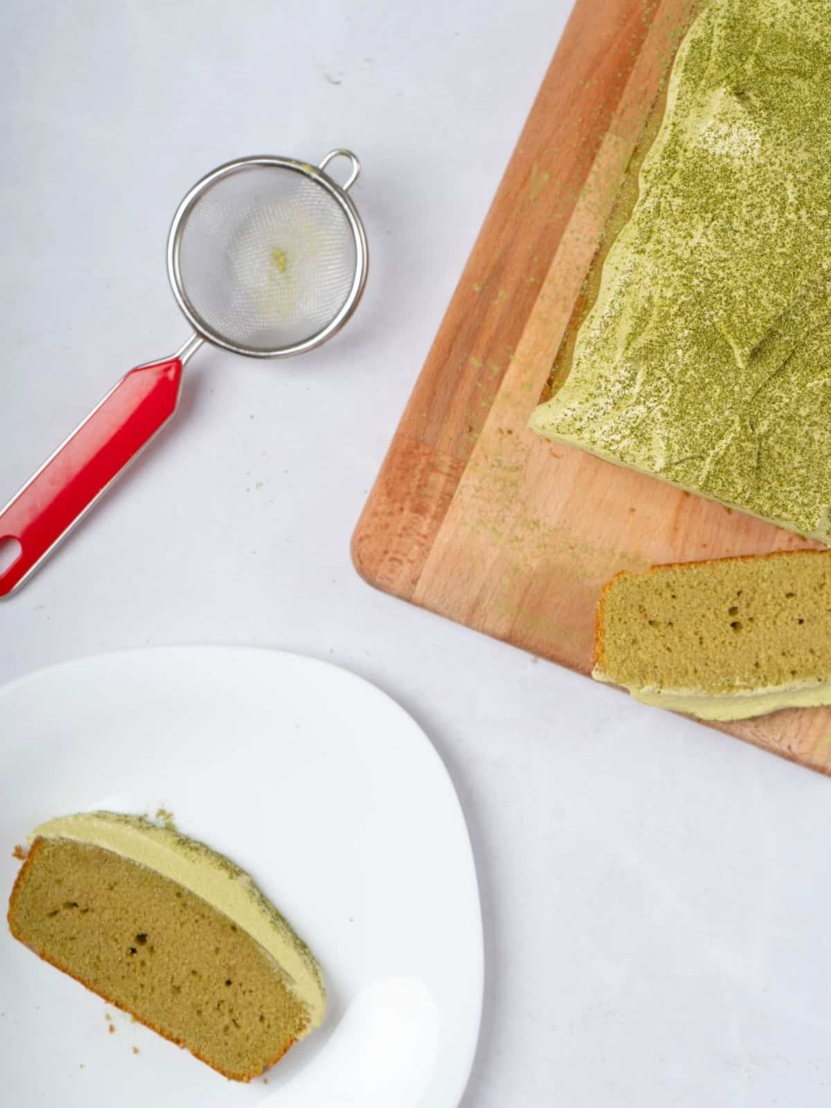 Matcha cake sprinkled with matcha powder next to a strainer and a slice of matcha cake in a plate.