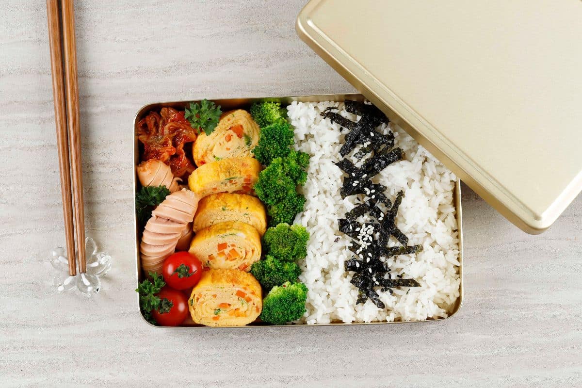 A traditional dosirak with rice, egg rolls, sausage, kimchi, and broccoli.