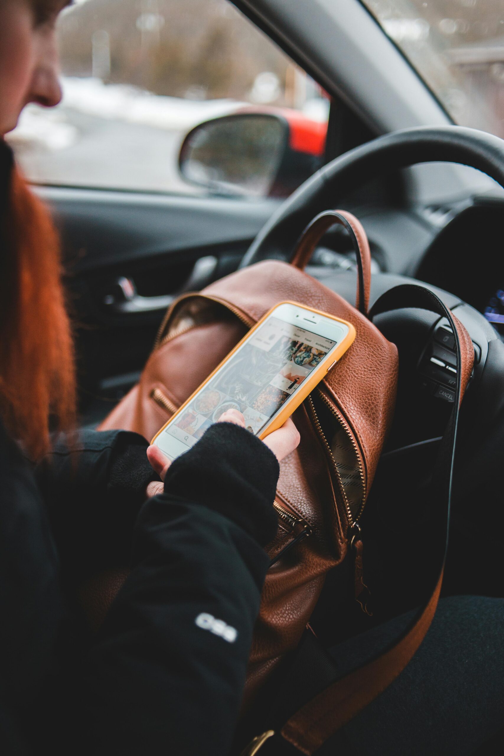 A person with red hair is sitting in a car, wearing a black coat and gloves, while holding a smartphone inside a bright yellow case. The phone screen shows an Instagram feed, and a brown leather bag rests on their lap.