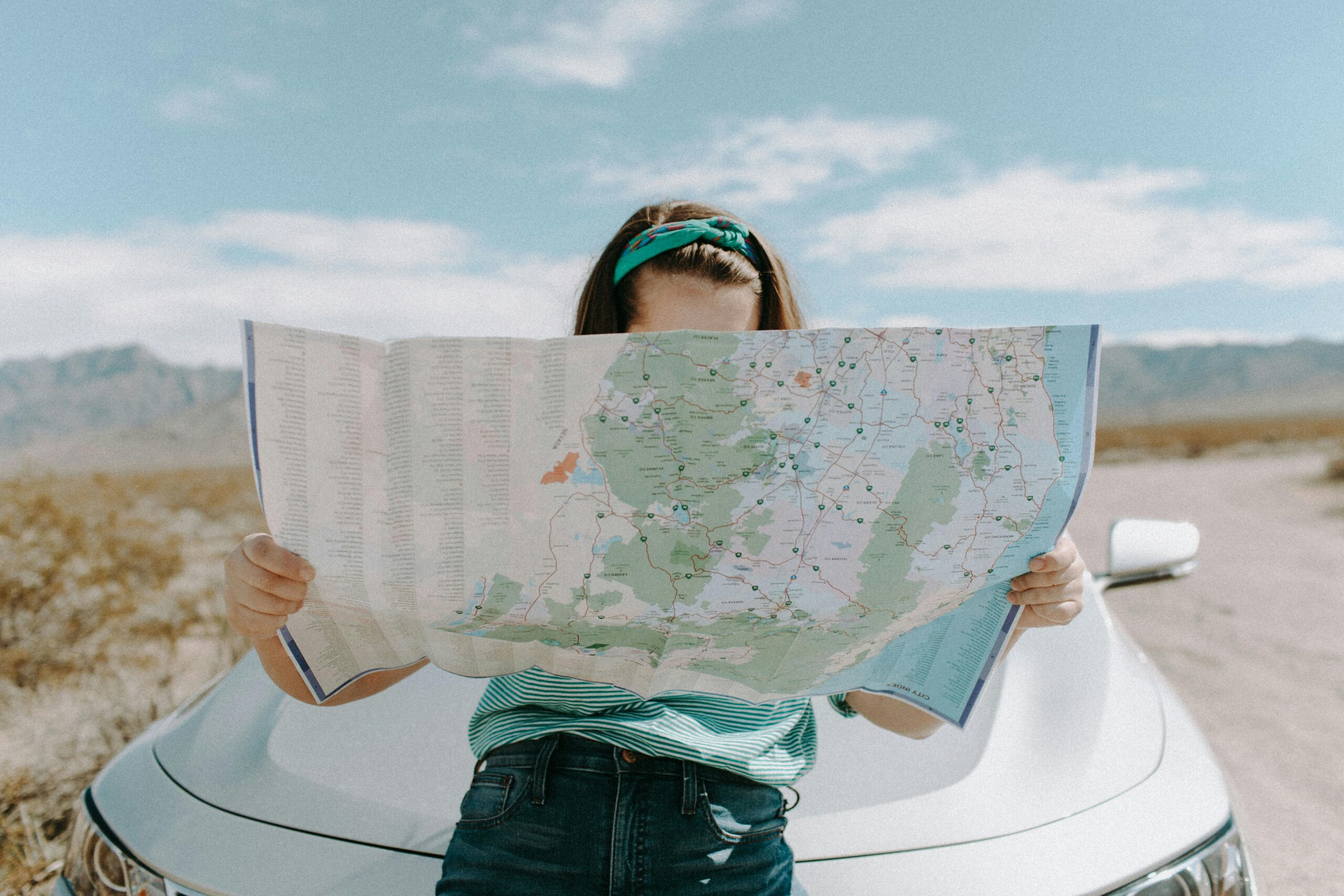 An image featuring a young woman standing in front of a car, holding open a large map that she is examining. The setting appears to be a dry landscape with mountains in the background. She is wearing a striped, green and white top and denim jeans. Her hair is styled back with a teal headband, adding a casual yet explorative touch to her outfit. 