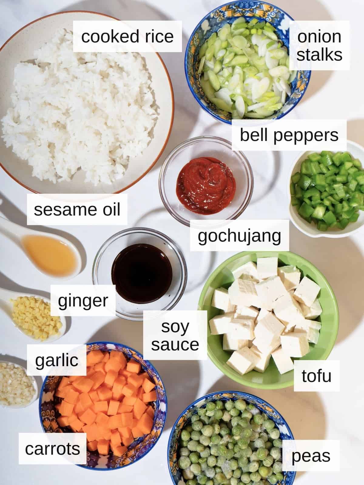 Ingredients for gochujang fried rice, including cooked rice, green onion stalks, bell peppers, sesame oil. ginger, garlic, carrots, soy sauce, tofu, peas, and gochujang.