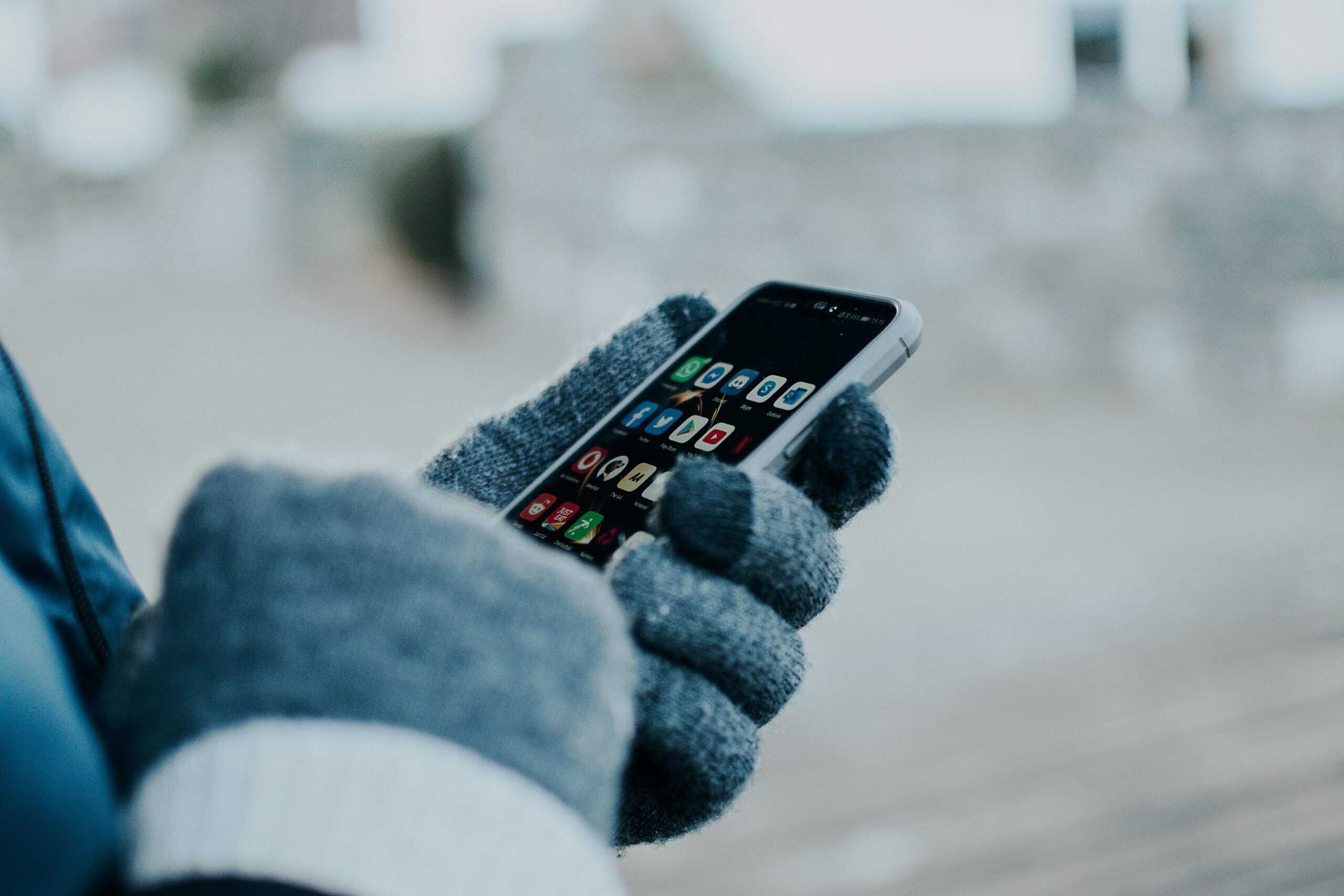 A person wearing a blue jacket and gray gloves is holding a smartphone outside in a wintry setting. The screen displays various app icons, suggesting a moment of connectivity or navigation outdoors.