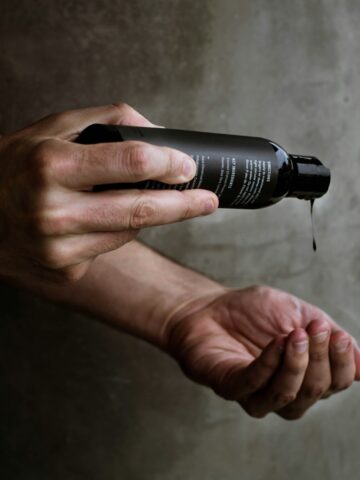 Close-up of a man’s hands pouring liquid skincare product from a matte black bottle against a gray concrete wall.