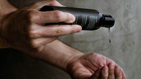 Close-up of a man’s hands pouring liquid skincare product from a matte black bottle against a gray concrete wall.