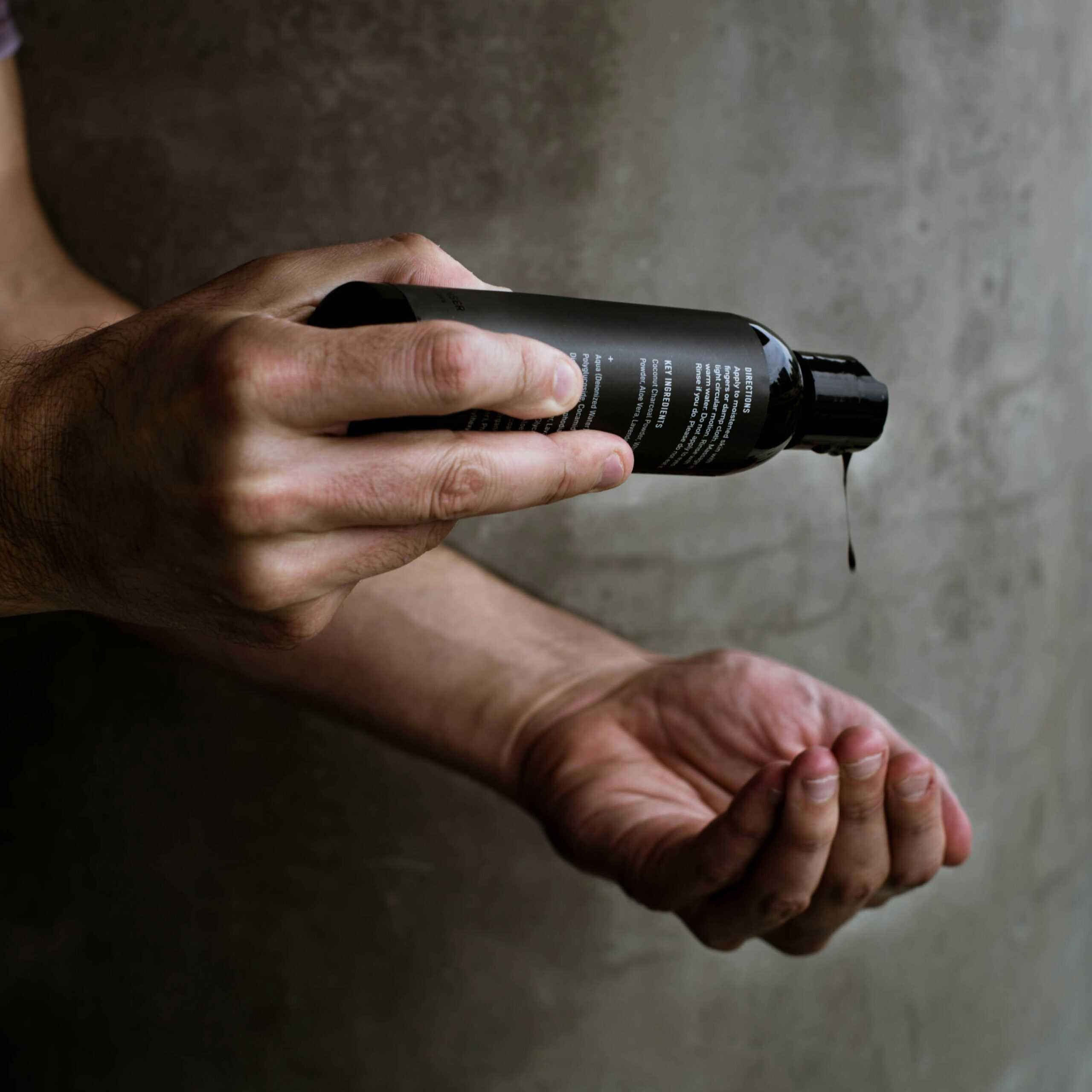 Close-up of a man’s hands pouring liquid skincare product from a matte black bottle against a gray concrete wall.