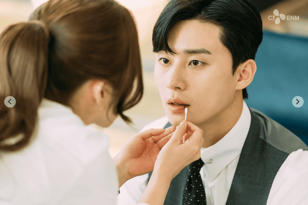 Close-up of Secretary Kim gently applying lip balm to Vice Chairman Lee Young-joon as he sits still, wide-eyed and stunned in his suit.