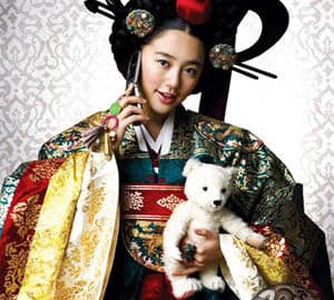 Actress in full royal Korean attire holding a white teddy bear and a cellphone, portraying a playful contrast between tradition and modern life.