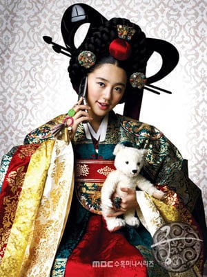 Actress in full royal Korean attire holding a white teddy bear and a cellphone, portraying a playful contrast between tradition and modern life.