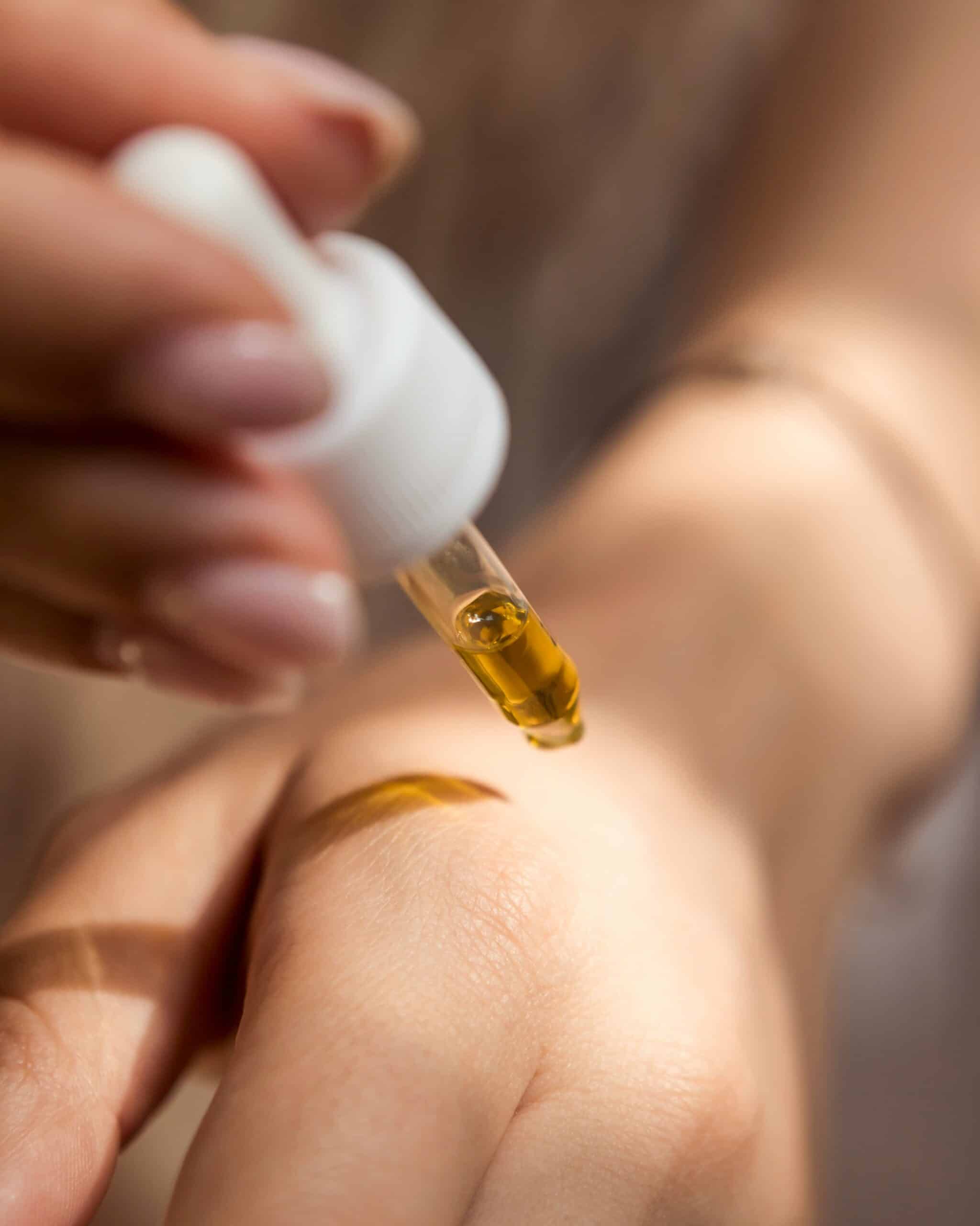 A dropper applying golden skincare oil onto the back of a hand, with a blurred nude-toned background.