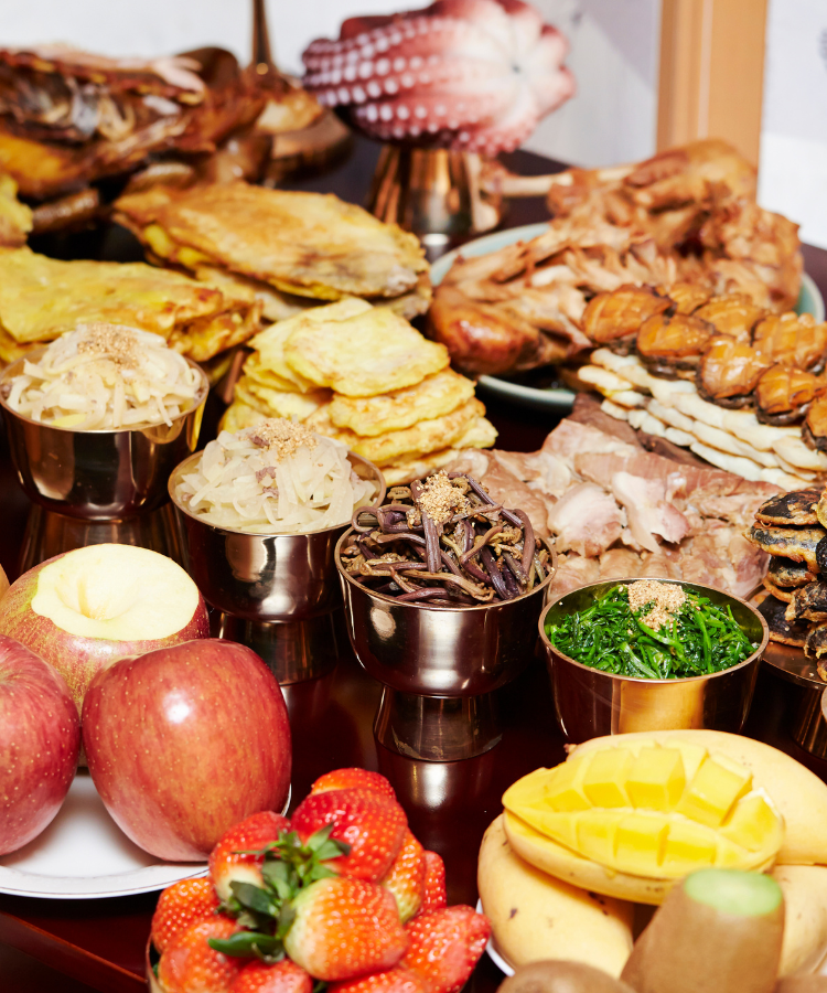 Close-up of a Chuseok feast featuring jeon, fresh fruits, dried fish, and traditional Korean side dishes in brass bowls.