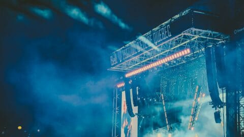 A wide view of a packed outdoor concert at night with blue lighting and a large stage setup.