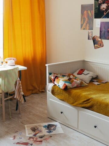 K-pop styled bedroom with mustard yellow curtain, cozy bed, and a small photocard gallery on the wall.