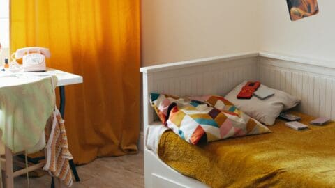K-pop styled bedroom with mustard yellow curtain, cozy bed, and a small photocard gallery on the wall.