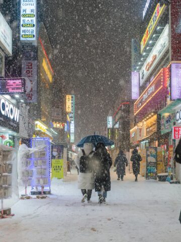 Snowy street scene in Seoul Korea with illuminated signs, people with umbrellas, vibrant city nightlife.