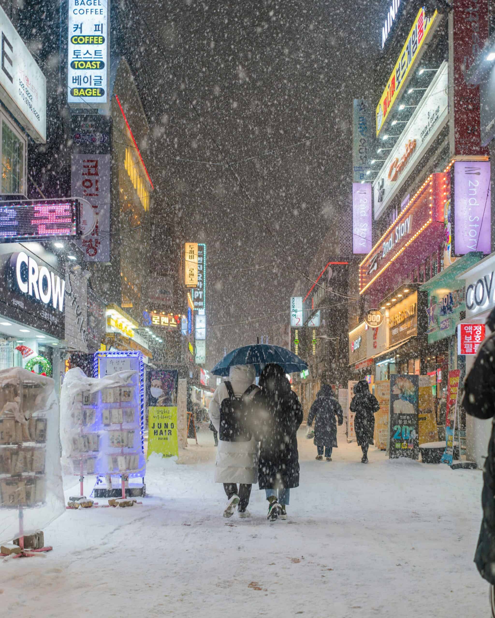 Snowy street scene in Seoul Korea with illuminated signs, people with umbrellas, vibrant city nightlife.