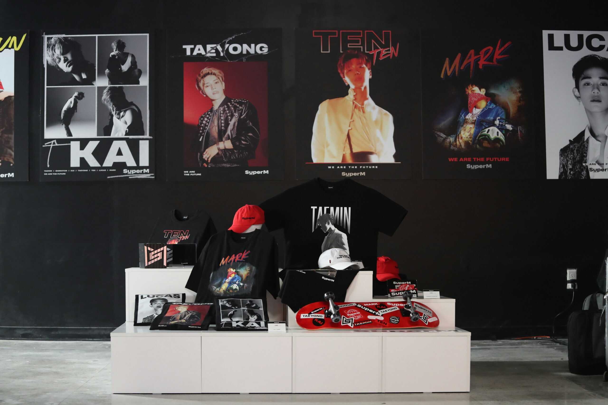 A K-pop merchandise display featuring SuperM member posters, branded shirts, hats, and albums on a white showcase stand.