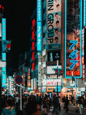 A busy night street in Tokyo filled with neon signs, billboards featuring music groups, and a crowd of pedestrians—capturing the vibrant energy of a city where pop culture thrives.