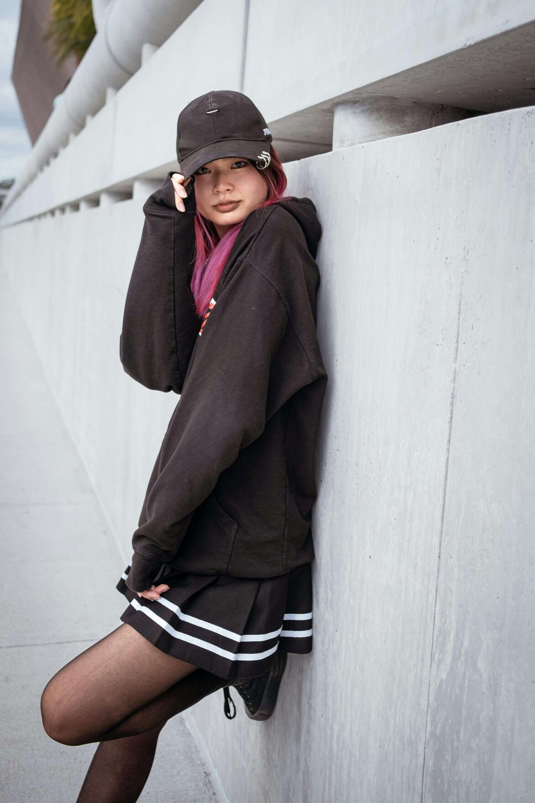 Stylish young woman in black hoodie, cap, with pink hair, urban backdrop in Seoul Korea.