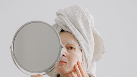 Woman in a towel wrap applying a sheet mask in front of a hand mirror